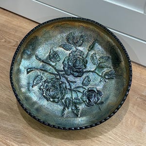 May include: An iridescent glass bowl with a raised floral design featuring roses and leaves. The bowl has a dark, textured rim and a shimmering, multi-toned surface, reflecting shades of blue, green, and gold.
