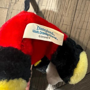 May include: A Mickey Mouse soft toy with a red head, black ears, and yellow feet. The toy has a tag that reads "Disneyland Walt Disney World &copy;DISNEY". The toy is made of soft, plush material.