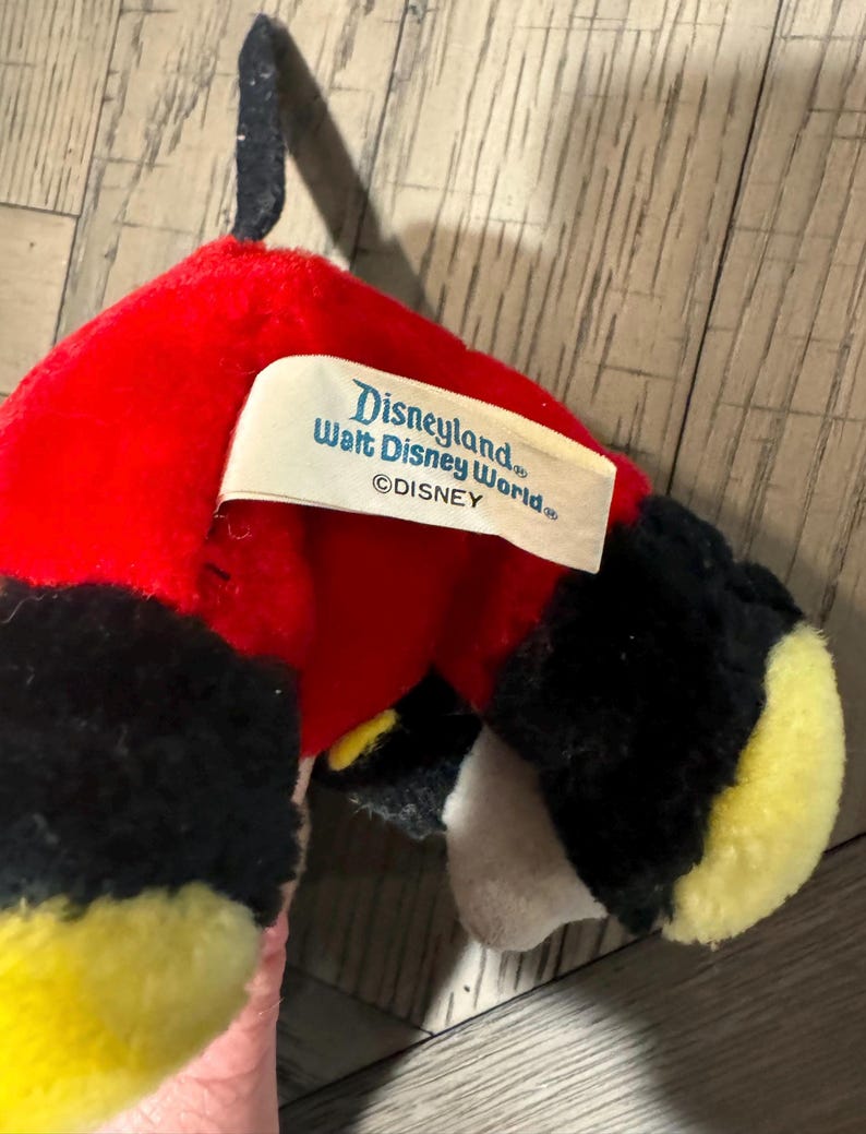 May include: A Mickey Mouse soft toy with a red head, black ears, and yellow feet. The toy has a tag that reads "Disneyland Walt Disney World &copy;DISNEY". The toy is made of soft, plush material.