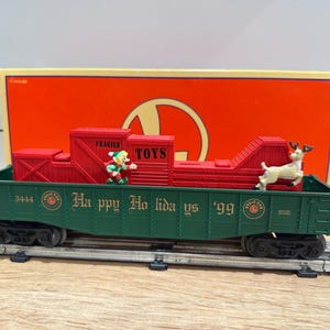May include: A green and red toy train car with the words "Happy Holidays '99" printed on the side. The train car has a red cargo area with the word "TOYS" and a small elf figurine. A reindeer figurine is also on the train car.