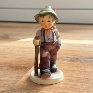 May include: A ceramic figurine of a young hiker wearing a green hat with a red feather, a light purple shirt, and brown trousers with suspenders. The figure holds a brown walking stick and stands on a circular white base, painted in muted tones.