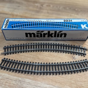 Puede incluir: Una caja Märklin azul y blanca con secciones de vía curva y dos vías de tren modelo curvas. La caja dice "Gebogene Gleisstücke" y "2231". Las vías son negras con rieles plateados.