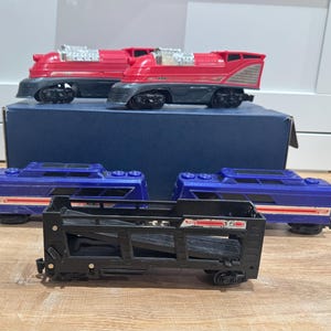 May include: A collection of toy train cars in red and blue. The red train cars are on top, and the blue ones are below. A black flatbed car is in the foreground. The toy trains are on a wooden surface.