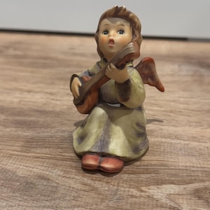 Puede incluir: Figura de cerámica de un ángel tocando la guitarra. El ángel tiene cabello castaño, una túnica verde claro y pequeñas alas rojas. La guitarra es marrón con una boca de sonido marrón claro. La figura está sobre una superficie de madera.