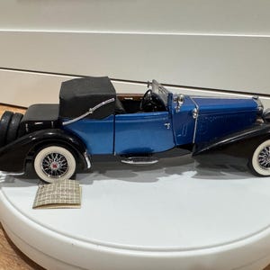 May include: A detailed model of a classic blue convertible car with black fenders and a black convertible top. The car has white-walled tires with red accents and a spare tire on the back. The model sits on a white surface.