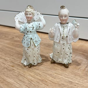 以下が含まれることがあります： 伝統的な衣装を着た女性の磁器製フィギュア2体。一方は水色のトップスを着て扇子を持ち、もう一方は白いジャケットを着て小さな物を持っています。どちらも顔の表情が細かく、金色のアクセントが施されています。