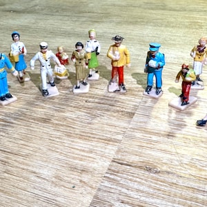 May include: A collection of miniature figurines depicting various professions and characters. The figurines are painted in bright colors and stand on small, rectangular bases. The collection includes figures in uniforms, casual wear, and carrying props.