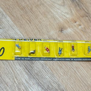 May include: A pack of HO scale model figures in various poses. The figures are hand-painted and come in individual clear plastic compartments. The packaging is yellow with black text and the word "ho" in black.