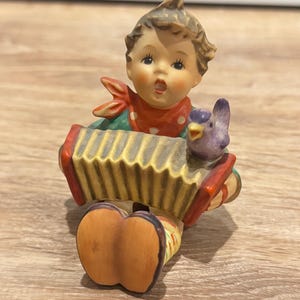May include: A ceramic figurine of a child playing an accordion. The figurine has a red and white polka dot scarf, green shirt, and brown hair. A purple bird sits on the accordion. The accordion is yellow and red.