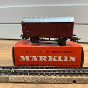 May include: A model train car, in maroon and silver, sits atop a red and orange box. The box reads "CLOSED GOODS VAN, WITH TAIL LIGHTS" and "MARKLIN". Model train tracks are in the foreground.