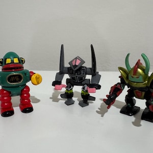 May include: Three colorful toy robots. The robot on the left is green, red, and yellow. The middle robot is gray and pink. The robot on the right is black, red, and gold. Each robot has a unique design.