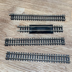May include: Four sections of model train tracks. The tracks are black with gold-colored rails. One section has a black plastic connector. The tracks are arranged on a light brown wooden surface.