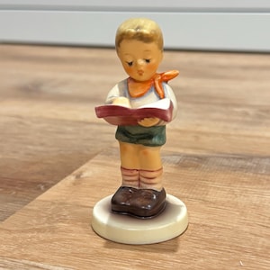 May include: A ceramic figurine of a young person with blonde hair, wearing a white shirt, orange neckerchief, and green shorts, standing on a circular base while holding an open red book with white pages and wearing brown shoes.