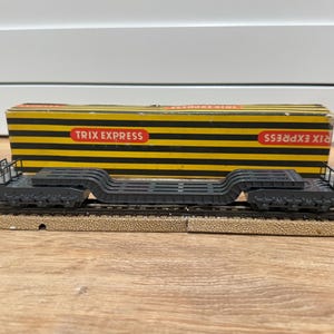 Puede incluir: Un vagón plano de tren modelo con una caja a rayas negras y amarillas con la etiqueta "TRIX EXPRESS". El vagón es de color gris oscuro y se encuentra sobre una sección de vías de tren modelo. La caja es rectangular con letras rojas.