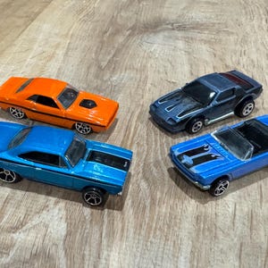 May include: Four toy cars are arranged on a wooden surface. The cars are in various colors, including orange, blue, and dark gray. One is a convertible. The cars have black wheels and detailed designs.