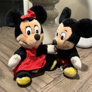 May include: Two plush Mickey and Minnie Mouse dolls. Minnie wears a red dress with white polka dots and a matching bow. Mickey is in black shorts, red trousers, and yellow shoes. Both have black ears and tan faces.