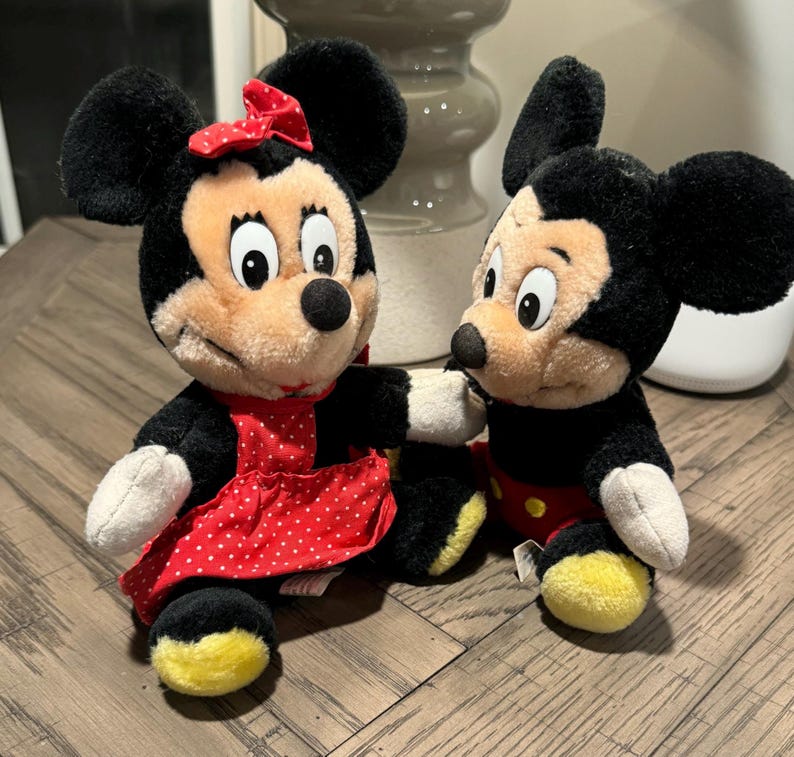 May include: Two plush Mickey and Minnie Mouse dolls. Minnie wears a red dress with white polka dots and a matching bow. Mickey is in black shorts, red trousers, and yellow shoes. Both have black ears and tan faces.