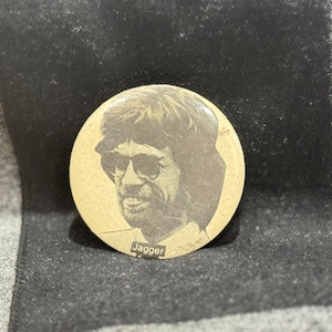 Könnte beinhalten: Ein runder, beigefarbener Knopf mit einem Sepia-Bild eines Mannes mit Sonnenbrille. Der Name "Jagger" ist unten in Weiß aufgedruckt. Der Knopf liegt vor einem dunklen Hintergrund.