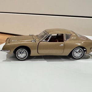 May include: A gold-colored die-cast model car with a classic design. The car has a sleek, aerodynamic shape, chrome accents, and white-walled tires. The model is displayed on a white surface.
