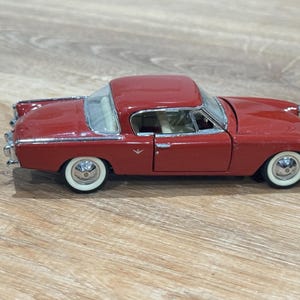 May include: A red vintage toy car with white-walled tires. The classic car has a detailed chrome trim, a white interior, and a clear windshield. The car is a miniature replica of a classic automobile.