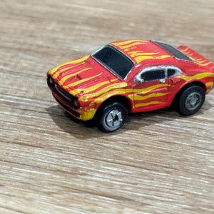 May include: A small, red toy car with yellow flame decals on the hood and sides. The car has black windows and silver wheels. The toy car is a miniature replica of a classic sports car.