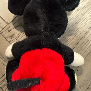 May include: A plush Mickey Mouse toy, seen from the rear. The toy has black fur, large round ears, white gloves, and red shorts. The toy is lying on a wooden surface.