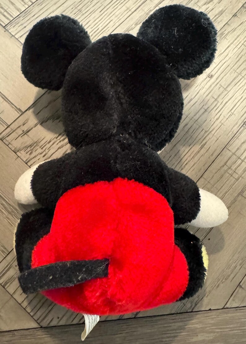 May include: A plush Mickey Mouse toy, seen from the rear. The toy has black fur, large round ears, white gloves, and red shorts. The toy is lying on a wooden surface.