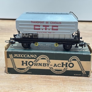 May include: A model train car labeled "TRANSPORT DE CEREALES C.T.C" in red lettering. The train car is gray and sits on a black chassis with black wheels. It rests on a green and brown box with "MECCANO HORNBY-ACHO PARIS" printed on it.