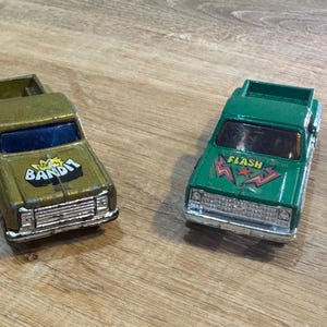 Puede incluir: Dos camionetas de juguete. Una es de color verde oliva con el texto "Bandit", la otra es verde con el texto "Flash". Ambas tienen parachoques y parabrisas plateados. Los juguetes están sobre una superficie de madera.