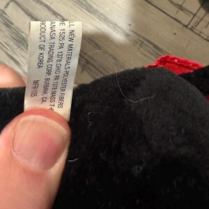 May include: A close-up of a black plush toy with a red accent. A white tag is visible, with text including "LL NEW MATERIALS-POLYESTER FIBERS" and "ANASA TRADING CORP. BURBANK, CA."