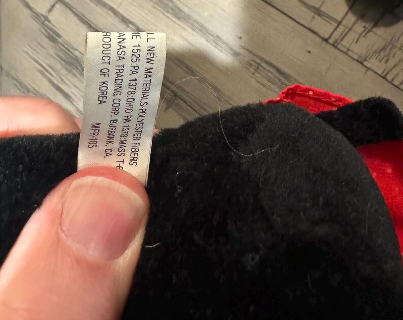 May include: A close-up of a black plush toy with a red accent. A white tag is visible, with text including "LL NEW MATERIALS-POLYESTER FIBERS" and "ANASA TRADING CORP. BURBANK, CA."