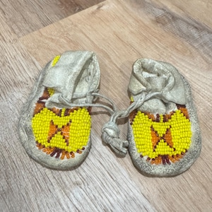 May include: A pair of baby moccasins with a beaded design. The moccasins are made of a light gray material and feature a yellow and orange beaded pattern on the toe. The laces are tied together.