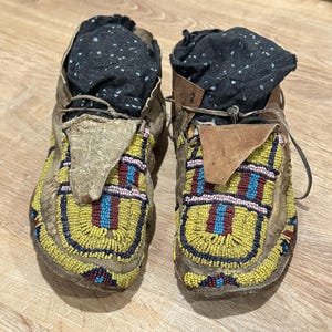 Puede incluir: Un par de mocasines con cuentas vintage. Los zapatos están hechos de cuero desgastado y presentan intrincados abalorios en amarillo, rojo y azul. La parte superior de los zapatos está rellena de tela negra. Los mocasines están atados con cordones de cuero.
