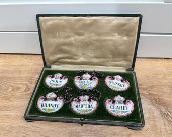 May include: An antique set of six decorative liquor labels, each with a floral border and a different liquor name: Port, Liqueur Brandy, Whiskey, Brandy, Madeira, and Claret. The labels are displayed in a vintage green velvet-lined box.