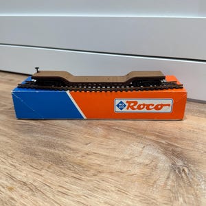 May include: A brown model railway flatcar sits on a short section of black track, which is placed atop a rectangular box. The box features a blue and orange colour scheme with the white and blue Roco logo printed on the front side.