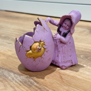 May include: A lavender-colored decorative figurine featuring a child in a bonnet standing beside a large, cracked egg. A yellow chick is emerging from the center of the egg, which has gold-painted edges along the cracks and a textured surface.