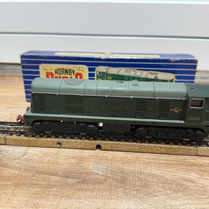 Könnte beinhalten: Eine grüne und graue Modell-Lokomotive auf einer Schiene, mit der Nummer 08000 sichtbar. Eine blaue Schachtel mit dem Wort "HORNBY" befindet sich im Hintergrund. Das Modell ist eine detaillierte Nachbildung.
