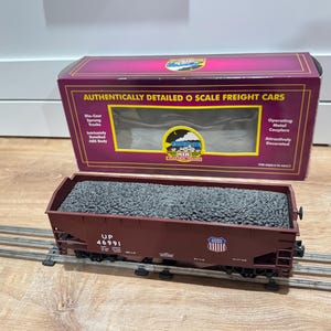 May include: A maroon O scale freight car model filled with gray material, sitting on a model train track. The box behind it reads "Authentically Detailed O Scale Freight Cars". The car has the number "UP 46991" printed on the side.