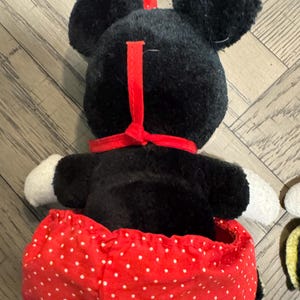 May include: A plush Minnie Mouse doll, seen from the back, with a black head, large ears, a red bow, and a red and white polka dot skirt. The doll has white gloves and a red collar.