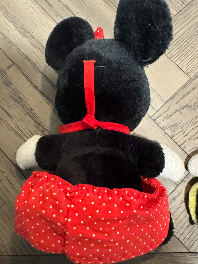 May include: A plush Minnie Mouse doll, seen from the back, with a black head, large ears, a red bow, and a red and white polka dot skirt. The doll has white gloves and a red collar.