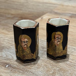 Puede incluir: Dos vasos de chupito hexagonales negros con ilustraciones doradas y beige de una figura sentada. Los vasos tienen un halo dorado y un interior beige. El fondo es negro.