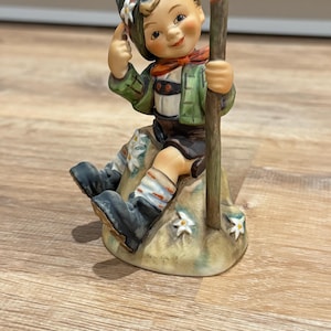 Könnte beinhalten: Keramikfigur eines lächelnden Jungen in traditioneller bayerischer Tracht. Er trägt eine grüne Jacke, braune Shorts und schwarze Stiefel. Der Junge sitzt auf einem Felsen, hält einen Holzstab und trägt einen Hut mit einer Blume.