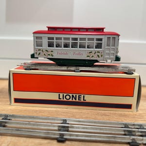 May include: A vintage model train trolley car with a red roof and white body, labeled "Yuletide Trolley." It sits atop a box with the "Lionel" logo, with train tracks in the foreground. The trolley has decorative holiday accents.