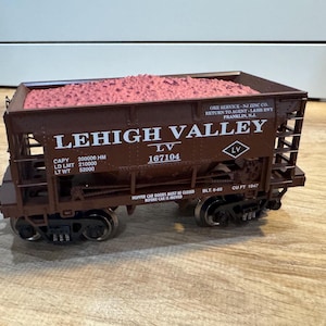 May include: A brown model train car from the Lehigh Valley railroad, filled with pink material. The car has the text "LEHIGH VALLEY" and the number "167104" printed on the side. The car is on a wooden surface.