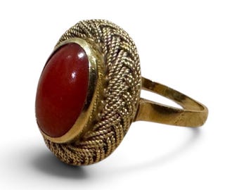 May include: A gold ring featuring a smooth, oval-shaped red stone set in the center. The stone is surrounded by a textured, braided gold metal border that creates a wide, circular frame. The band of the ring is a simple, plain gold metal.