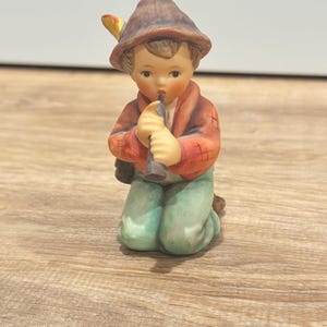 Puede incluir: Una figura de cerámica de un niño arrodillado tocando la flauta. El niño lleva un sombrero marrón con una pluma, una chaqueta roja y pantalones verdes. La figura tiene una apariencia detallada, pintada a mano, con una estética clásica y vintage.
