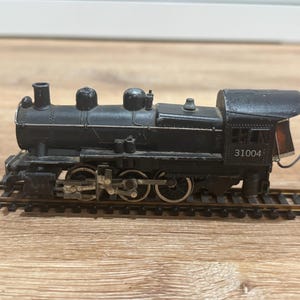 Puede incluir: Una locomotora de tren modelo negra con el número 31004 en el lateral. El tren está sobre una vía marrón y tiene varias ruedas. La locomotora tiene una chimenea y otros detalles.