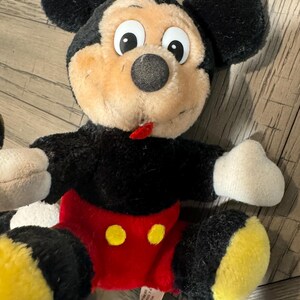 May include: A plush Mickey Mouse doll with black fur, a tan face, and large white-eyed features. The doll wears red shorts with yellow buttons and yellow feet. The doll has black ears and white gloves.