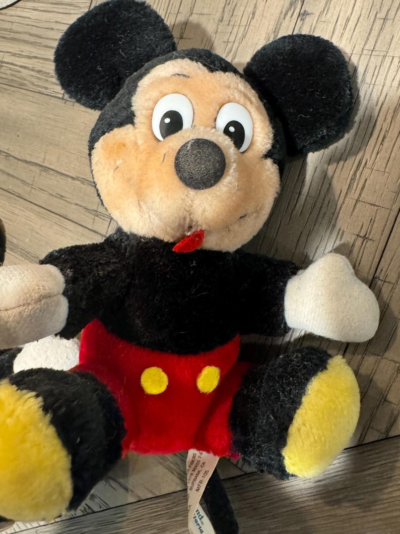 May include: A plush Mickey Mouse doll with black fur, a tan face, and large white-eyed features. The doll wears red shorts with yellow buttons and yellow feet. The doll has black ears and white gloves.