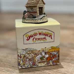 May include: A miniature figurine of a small house with a brown shingled roof and a wooden door, set atop a box. The box features the text "David Winter Cameos" and a detailed illustration of a village scene.